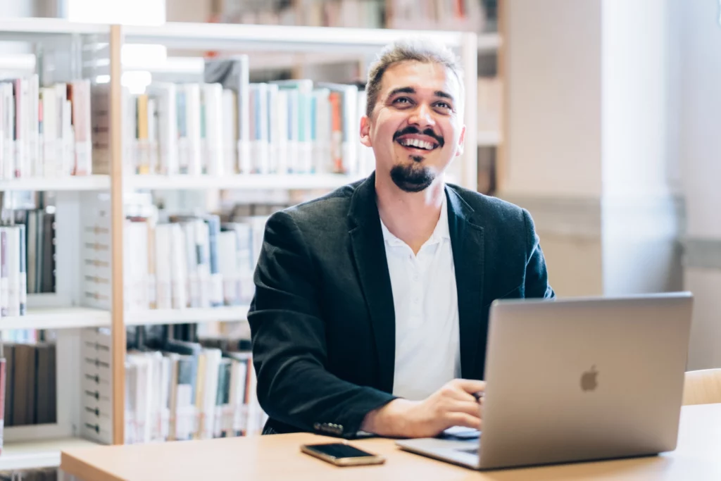 Kristian Selimi al lavoro con un laptop in una libreria, sorridente, in abito professionale.