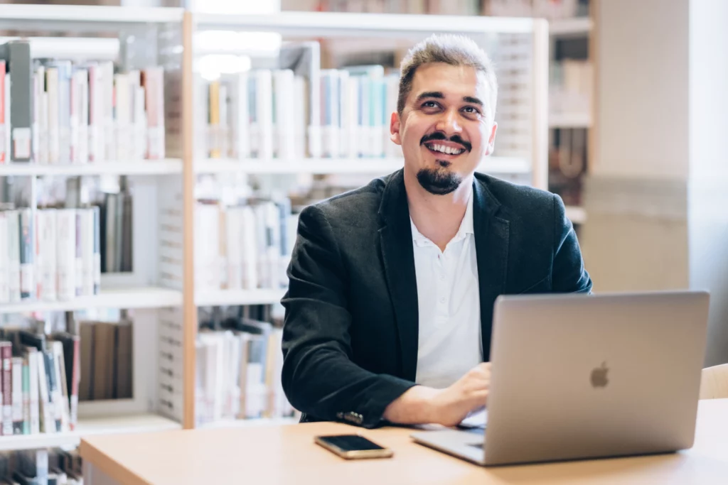 Kristian Selimi al lavoro con un laptop in una libreria, sorridente, in abito professionale.